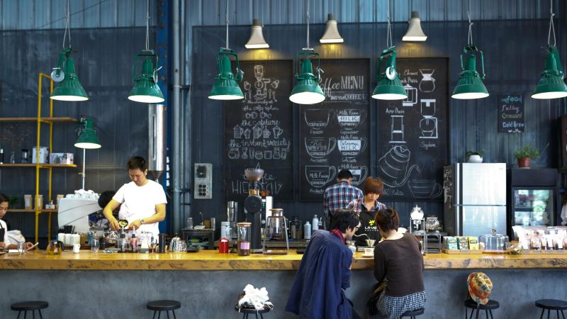 A bustling café with a modern industrial design, featuring a barista making coffee and patrons enjoying drinks.