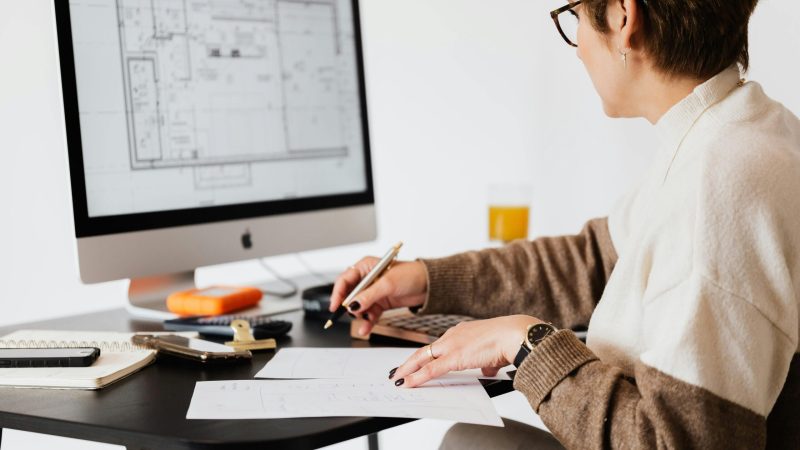 Woman designing architectural blueprints using desktop computer in a modern office setting.